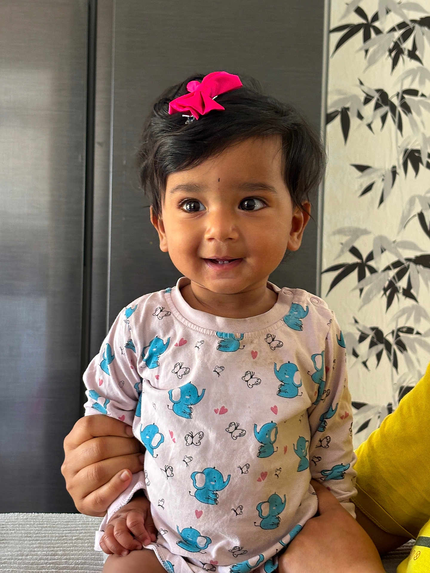 Mokshith joined the competition — help win amazing prizes! toddler, child, smiling, pink_bow, elephant_print, long_sleeve_shirt, hand, indoor, portrait, happy, cute, baby, person, patterned_clothing, holding, sitting, face, black_hair, wallpaper, yellow_sleeve
