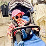 toddler, stroller, pink_cap, sunglasses, blue_cushion, black_socks, floral_shirt, shopping_store, clothing_rack, hanger, casual_clothes, child, indoor, person, footwear, seat, baby, retail, fashion, accessory