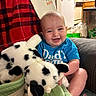 baby, smiling, blue_shirt, plush_toy, stuffed_animal, couch, blanket, indoor, kitchen, refrigerator, cabinet, basket, child, happy, person, seated, toy, home, cute, infant