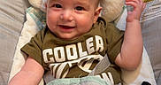 Ashton is registered to the contest to win money with this photo: baby, infant, child, smiling, face, green_clothing, camouflage_pants, baby_seat, cushion, elephant_design, indoor, carpet, happy, cute, comfort, person, portrait, young_child, clothing, seatbelt