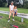 adult, baseball, baseball_bat, cap, casual_clothing, child, daylight, family, grass, guardrail, nature, outdoor, park, portrait, shorts, sitting, smiling, sneakers, sports, trees
