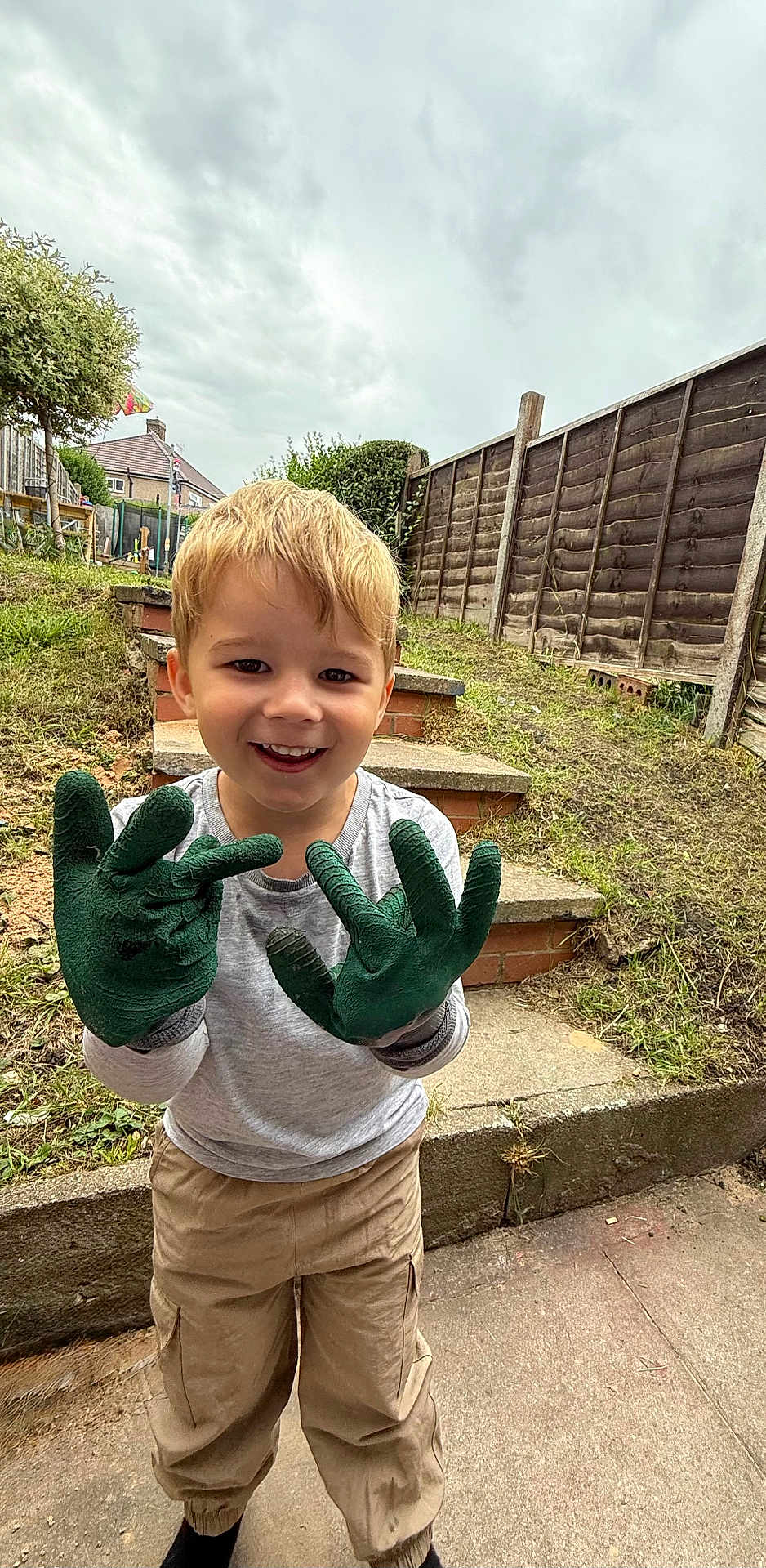 Logan joined the competition — help win amazing prizes! child, boy, smiling, gloves, green_gloves, gardening, outdoor, backyard, steps, fence, grass, cloudy_sky, pants, long_sleeve_shirt, happy, person, young_child, playful, casual_clothing, daytime