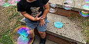 Logan is registered to the contest to win money with this photo: child, boy, bubbles, outdoor, steps, concrete, grass, black_tshirt, shorts, playful, smiling, happy, toy, bubble_container, sidewalk, casual_clothing, daylight, nature, fun