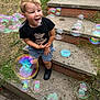 child, boy, bubbles, outdoor, steps, concrete, grass, black_tshirt, shorts, playful, smiling, happy, toy, bubble_container, sidewalk, casual_clothing, daylight, nature, fun