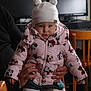 Jade a rejoint le concours — aidez-le/la à gagner de superbes lots ! adult_hands, baby, cartoon_print, child, clothing, computer_monitor, cozy, cute, expression, indoor, person, pink_jacket, pom_poms, portrait, puffy_jacket, seated, toddler, warm_lighting, white_hat, wooden_chair