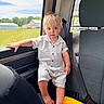 toddler, child, car_interior, seat, window, striped_clothing, barefoot, blonde_hair, blue_eyes, yellow_towel, orange_ring, daylight, green_fields, car_seatbelt, portrait, sitting, young_child, casual_clothing, curious_expression, backseat