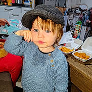 Timaël a rejoint le concours — aidez-le/la à gagner de superbes lots ! child, toddler, hat, blue_sweater, green_eyes, face, portrait, indoor, kitchen, food, fries, chicken_nuggets, takeout_container, wooden_table, messy_background, hand, candid, cap, snack, dining