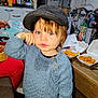child, toddler, hat, blue_sweater, green_eyes, face, portrait, indoor, kitchen, food, fries, chicken_nuggets, takeout_container, wooden_table, messy_background, hand, candid, cap, snack, dining