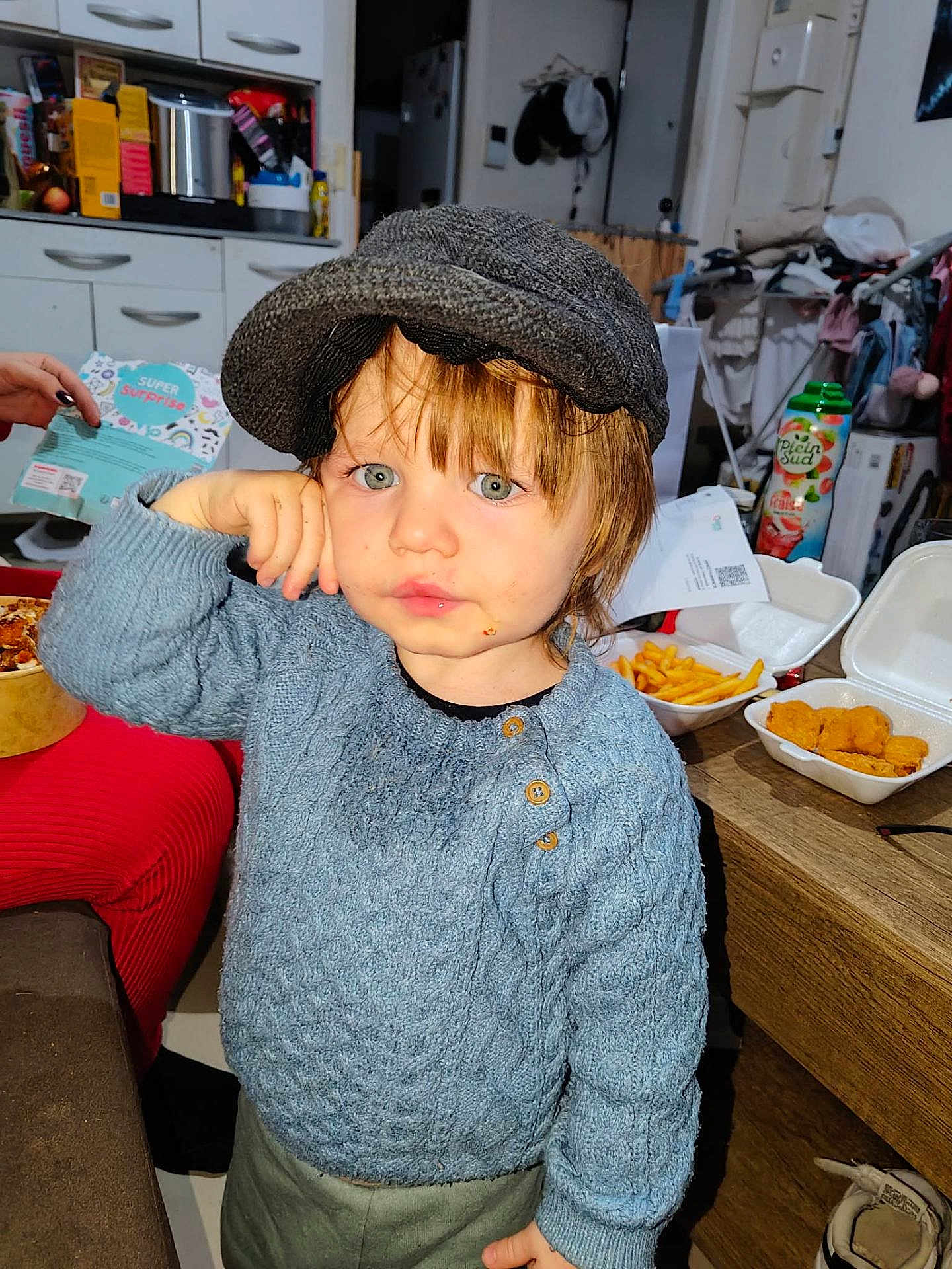Timaël a rejoint le concours — aidez-le/la à gagner de superbes lots ! child, toddler, hat, blue_sweater, green_eyes, face, portrait, indoor, kitchen, food, fries, chicken_nuggets, takeout_container, wooden_table, messy_background, hand, candid, cap, snack, dining