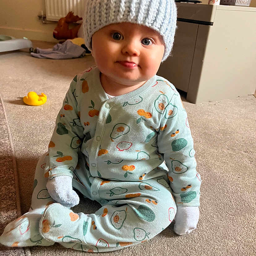 Denny James joined the competition — help win amazing prizes! baby, carpet, child, cozy, cute, domestic, expression, face, hat, home, indoor, infant, knit_hat, onesie, patterned_clothing, rubber_duck, sitting, soft_light, tongue_out, toy