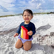 Mona joined the competition — help win amazing prizes! toddler, child, smiling, sand, beach, rainbow_shirt, purple_bottoms, kneeling, outdoor, sunny, clouds, sky, happy, cute, portrait, vacation, summer, playful, nature, daylight