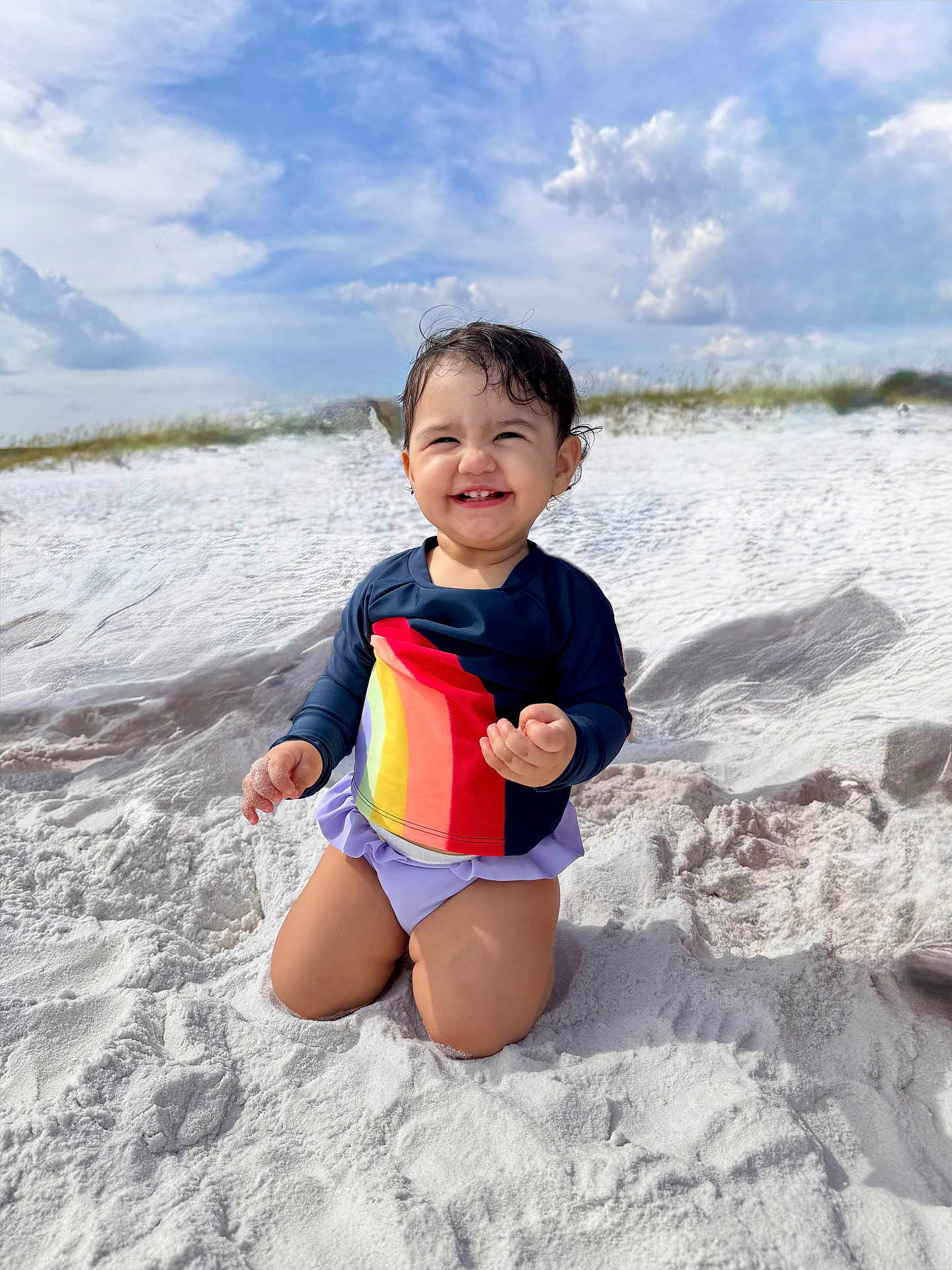 Mona joined the competition — help win amazing prizes! toddler, child, smiling, sand, beach, rainbow_shirt, purple_bottoms, kneeling, outdoor, sunny, clouds, sky, happy, cute, portrait, vacation, summer, playful, nature, daylight