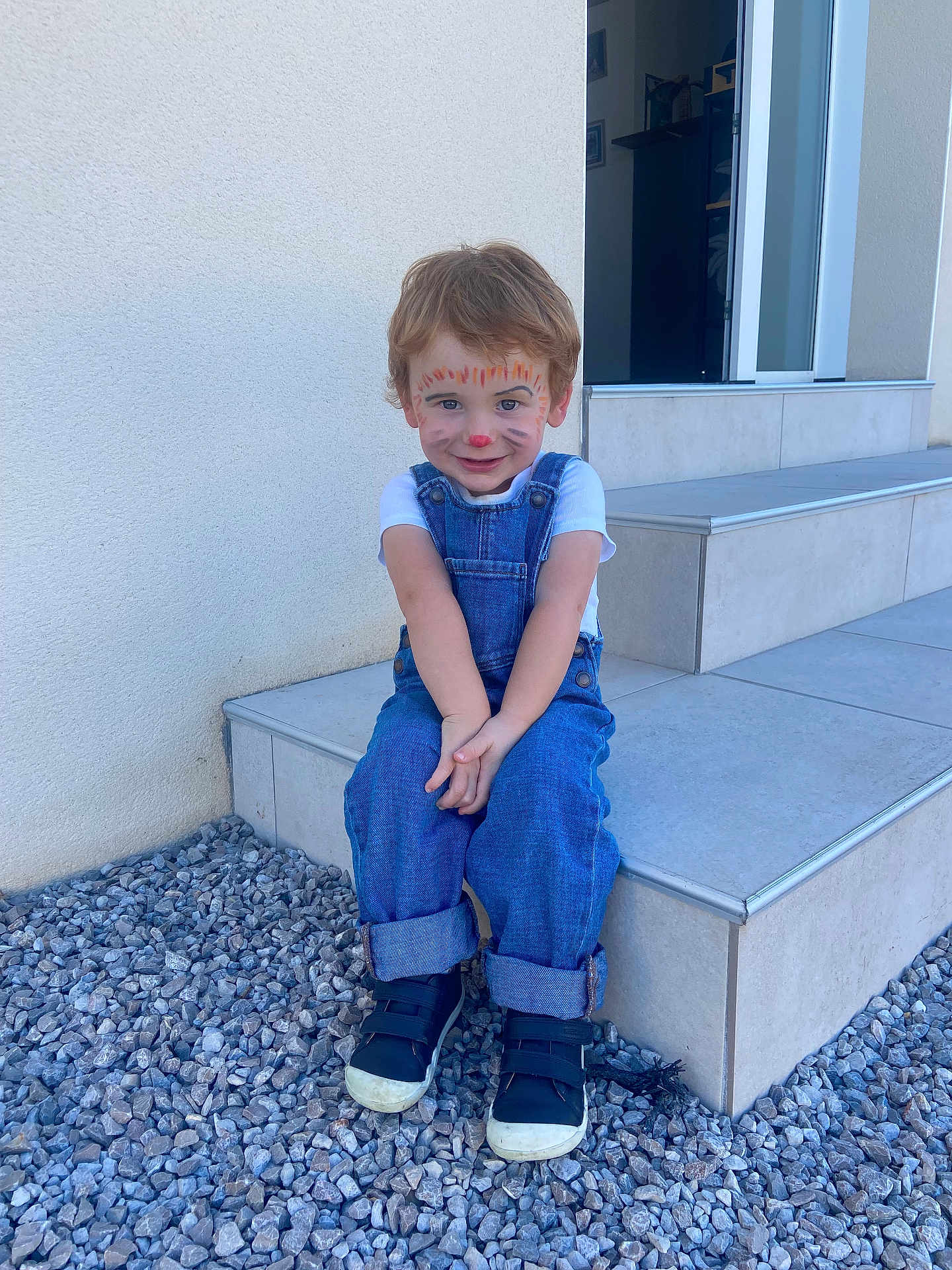 Sasha a rejoint le concours — aidez-le/la à gagner de superbes lots ! child, face_paint, tiger_face, denim_overalls, white_tshirt, black_sneakers, gravel, steps, outdoor, smiling, cute, young_child, casual_clothing, sitting, playful, daylight, curly_hair, wall, portrait, happy