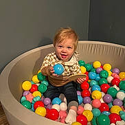 River joined the competition — help win amazing prizes! toddler, child, ball_pit, colorful_balls, indoor, play, toy, smile, happy, casual_clothing, sock, wooden_toy, floor, round_pit, blonde_hair, person, young_child, fun, playroom, curious