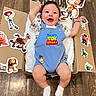baby, child, smiling, toy_story, romper, blue_clothing, cardboard_box, stickers, wooden_floor, indoor, happy, cute, infant, person, playful, decorations, blanket, sitting, smile, young_child