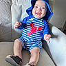 Yeshúa is registered to the contest to win money with this photo: baby, child, smile, blue_hat, striped_outfit, lobster_design, armchair, beige, indoor, happy, person, footwear, cute, infant, seated, portrait, clothing, texture, light, comfortable