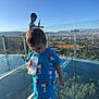 child, boy, baby_bottle, blue_clothing, snoopy, glass_floor, observation_deck, cityscape, mountains, trees, daylight, outdoor, sky, shorts, shoes, plastic_covers, glass_railing, person, playing, technology