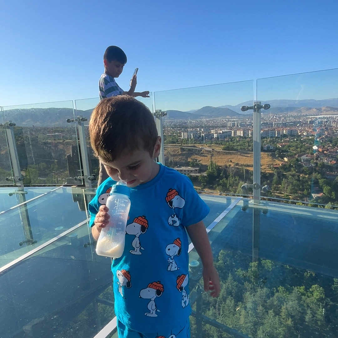 Hasan Emir a rejoint le concours — aidez-le/la à gagner de superbes lots ! baby_bottle, blue_clothing, boy, child, cityscape, daylight, glass_floor, glass_railing, mountains, observation_deck, outdoor, person, plastic_covers, playing, shoes, shorts, sky, snoopy, technology, trees