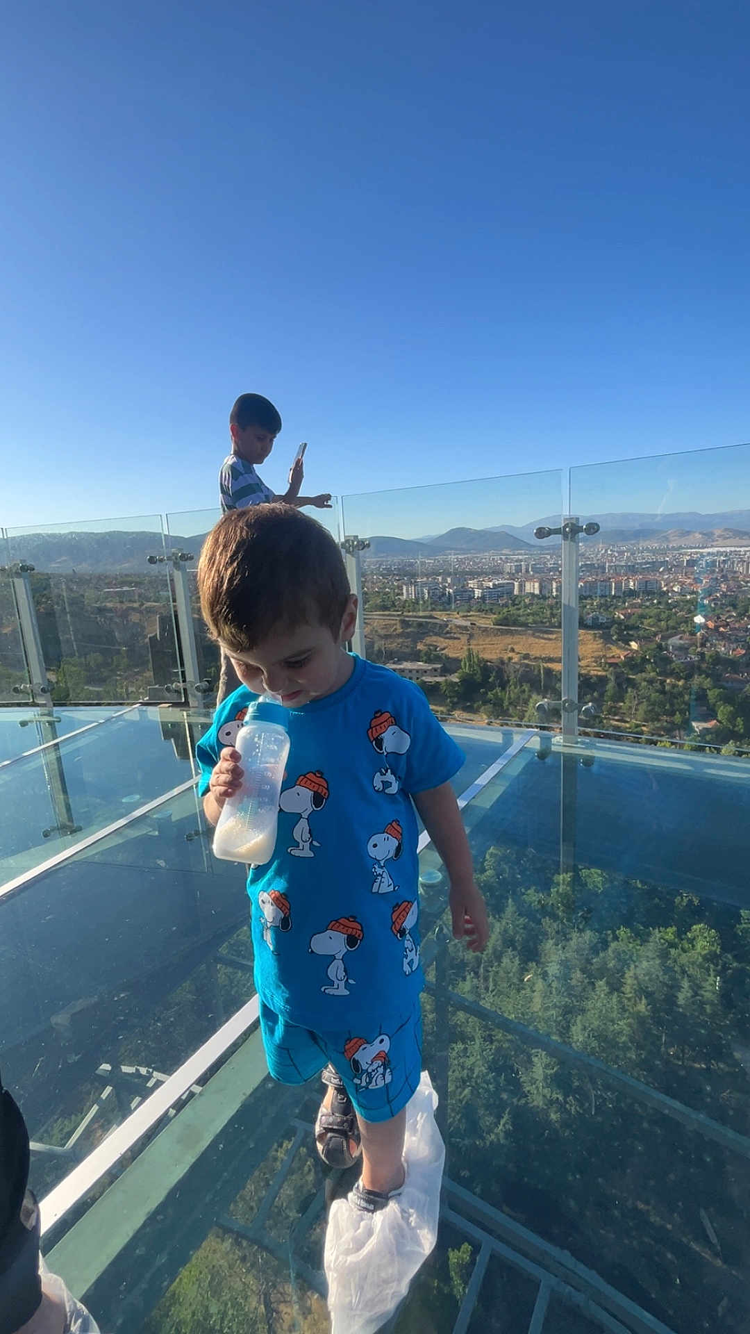 Hasan Emir a rejoint le concours — aidez-le/la à gagner de superbes lots ! child, boy, baby_bottle, blue_clothing, snoopy, glass_floor, observation_deck, cityscape, mountains, trees, daylight, outdoor, sky, shorts, shoes, plastic_covers, glass_railing, person, playing, technology