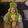 baby, child, couch, cozy, cute, dinosaur_costume, face, green_clothing, happy, headwear, home, indoor, infant, inflatable_chair, onesie, plush, portrait, seated, smiling, teddy_bear_prints
