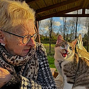 Lucky a rejoint le concours — aidez-le/la à gagner de superbes lots ! elderly_woman, cat, glasses, scarf, patterned_coat, tabby_cat, outdoor, wooden_roof, gazebo, trees, grass, sunlight, golden_hour, pet_collar, expressive_cat, companionship, portrait, cozy, bench, earrings