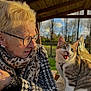 elderly_woman, cat, glasses, scarf, patterned_coat, tabby_cat, outdoor, wooden_roof, gazebo, trees, grass, sunlight, golden_hour, pet_collar, expressive_cat, companionship, portrait, cozy, bench, earrings