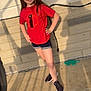 child, girl, smiling, red_tshirt, shorts, sneakers, outdoor, concrete, brick_wall, shadow, sunlight, posing, happy, casual_clothing, standing, playful, young, daylight, person, sidewalk