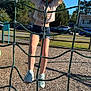 child, climbing, playground, rope_net, outdoor, summer, shorts, tshirt, sneakers, wood_chips, trees, cars, houses, blue_sky, daylight, person, recreation, fun, activity, park