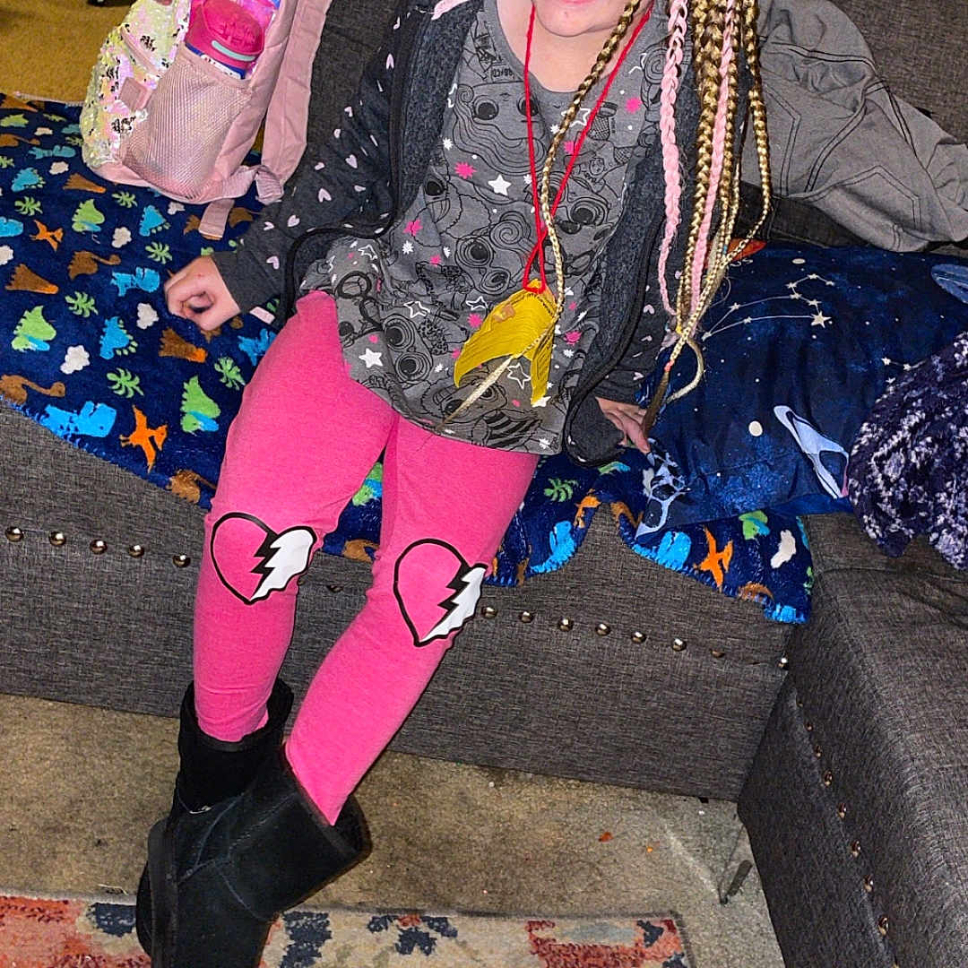 Niomi is registered to the contest to win money with this photo: backpack, black_boots, blanket, braided_hair, broken_heart, carpet, casual_clothing, child, colorful, couch, girl, happy, home, indoor, pillow, pink_leggings, portrait, seated, smiling, unicorn
