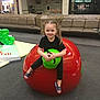 child, girl, smiling, pigtails, red_tomato, green_apple, mall, indoor, play_area, casual_clothing, slides, floor, bench, auntie_annes, pretzel_shop, toy, playground_equipment, carpet, happy, person