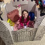 child, girl, cardboard_castle, pillow, toy, indoor, play, smiling, floor, patterned_pants, pink_shirt, backpack, clutter, carpet, seated, happy, imagination, home, casual, relaxed