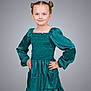 child, girl, portrait, smiling, hands_on_hips, teal_dress, long_sleeves, hair_buns, blue_eyes, standing, studio, gray_background, confident, cute, fashion, youth, person, pose, happy, casual