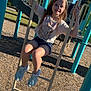 child, girl, playground, climbing, ladder, outdoor, daytime, smiling, casual_clothing, shorts, tshirt, sneakers, woodchips, play_equipment, metal_bars, trees, sky, sunlight, happy, recreation