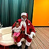 baby, child, santa_claus, adult, portrait, smiling, indoors, christmas, festive, red_suit, white_beard, santa_hat, couch, chair, curtain, orange_wall, carpet, family, holiday, photography