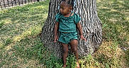 Siya is registered to the contest to win money with this photo: child, toddler, girl, outdoor, park, tree, grass, portrait, standing, leaning, green_outfit, braids, beads, hair_accessories, earrings, nature, sunlight, playground, fence, urban_park