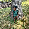 child, toddler, girl, outdoor, park, tree, grass, portrait, standing, leaning, green_outfit, braids, beads, hair_accessories, earrings, nature, sunlight, playground, fence, urban_park