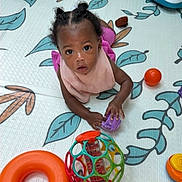 Cori is registered to the contest to win money with this photo: baby, child, infant, portrait, curious, looking_up, sitting, play_mat, toys, toy_ball, rattle, colorful, indoor, home, black_hair, braids, pink_clothing, smiling, floor, playtime