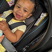 Kash is registered to the contest to win money with this photo: baby, child, car_seat, smile, curly_hair, bottle, hand, safety_harness, yellow_clothing, black_straps, infant, happy, portrait, indoors, seatbelt, person, cute, closeup, car_interior, joyful