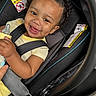 baby, child, car_seat, smile, curly_hair, bottle, hand, safety_harness, yellow_clothing, black_straps, infant, happy, portrait, indoors, seatbelt, person, cute, closeup, car_interior, joyful
