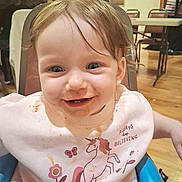 Romi joined the competition — help win amazing prizes! background, bib, chair, child, clothing, cute, face, hair, happy, high_chair, indoor, messy_face, people, person, sitting, smiling, table, toddler, unicorn, wooden_floor