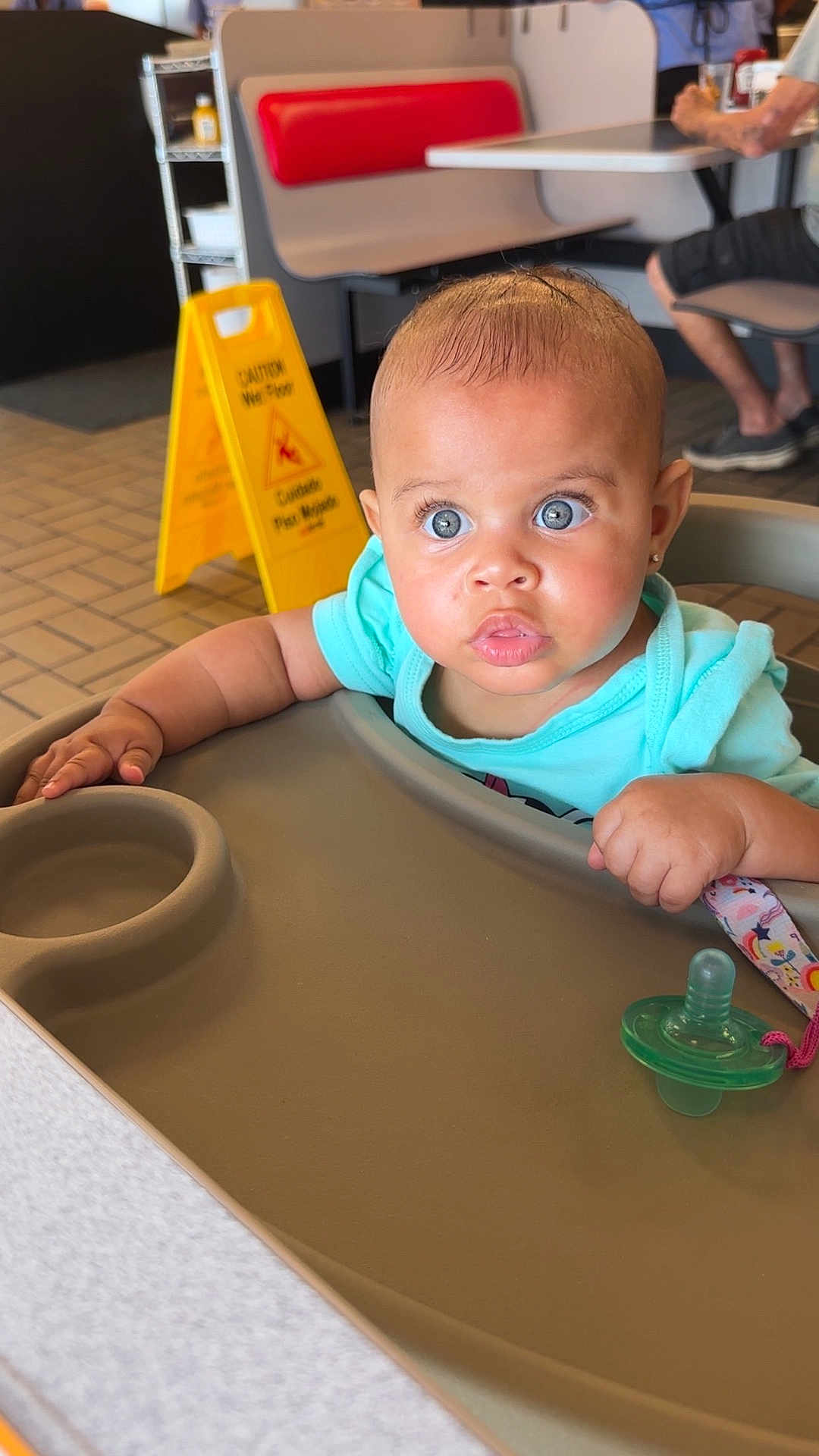 Leilanii joined the competition — help win amazing prizes! baby, high_chair, pacifier, blue_eyes, indoor, restaurant, table, floor_sign, caution_sign, child, person, skin, clothing, seat, hand, face, expression, furniture, people, casual