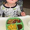 toddler, child, high_chair, pajamas, dinosaur_print, scrambled_eggs, toast, bacon, syrup, plate, green_plate, indoor, kitchen, food, meal, breakfast, person, blue_eyes, blonde_hair, expression