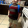 child, boy, portrait, standing, backpack, kitchen, stove, cereal_box, countertop, cabinets, sink, floor_tiles, striped_shirt, shorts, sneakers, indoors, home, looking_up, full_body, smiling