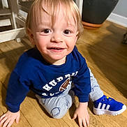 Mason is registered to the contest to win money with this photo: child, toddler, smiling, indoor, wooden_floor, blue_shirt, sneaker, left_hand, right_hand, brown_eyes, blonde_hair, plant_pot, pants, sitting, portrait, close_up, candid, floor, white_chair, happy