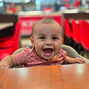 Leah joined the competition — help win amazing prizes! baby, child, smiling, high_chair, table, restaurant, indoor, red_chairs, happy, cute, person, infant, face, mouth_open, short_hair, clothing, seat, wood, blurred_background, bright_lighting