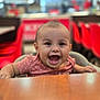 baby, child, smiling, high_chair, table, restaurant, indoor, red_chairs, happy, cute, person, infant, face, mouth_open, short_hair, clothing, seat, wood, blurred_background, bright_lighting