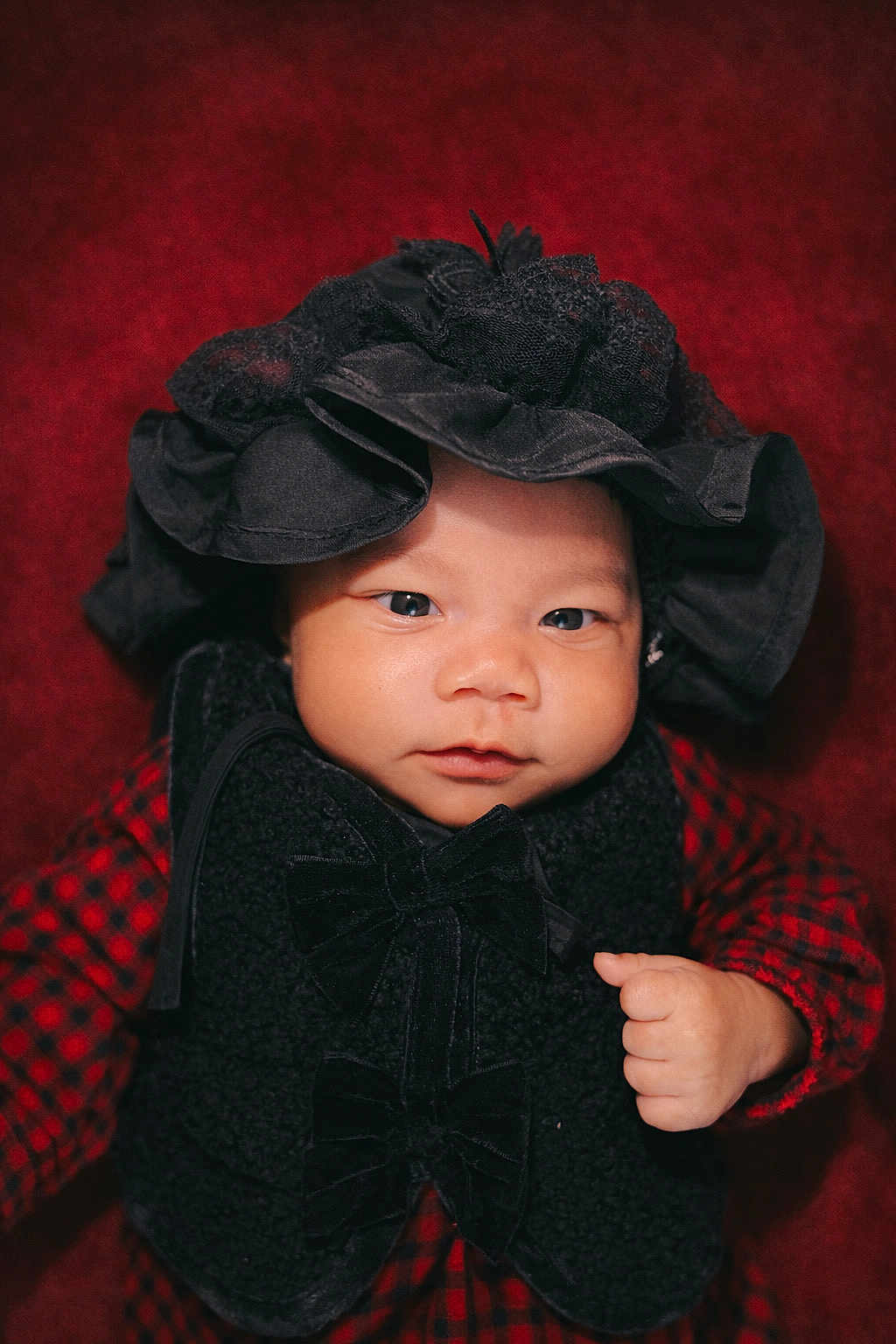 Hara-Mia joined the competition — help win amazing prizes! baby, infant, face, hat, black_hat, vest, black_vest, bow, red_checkered_clothing, hand, fist, portrait, closeup, cute, child, young, indoor, expression, looking_at_camera, red_background