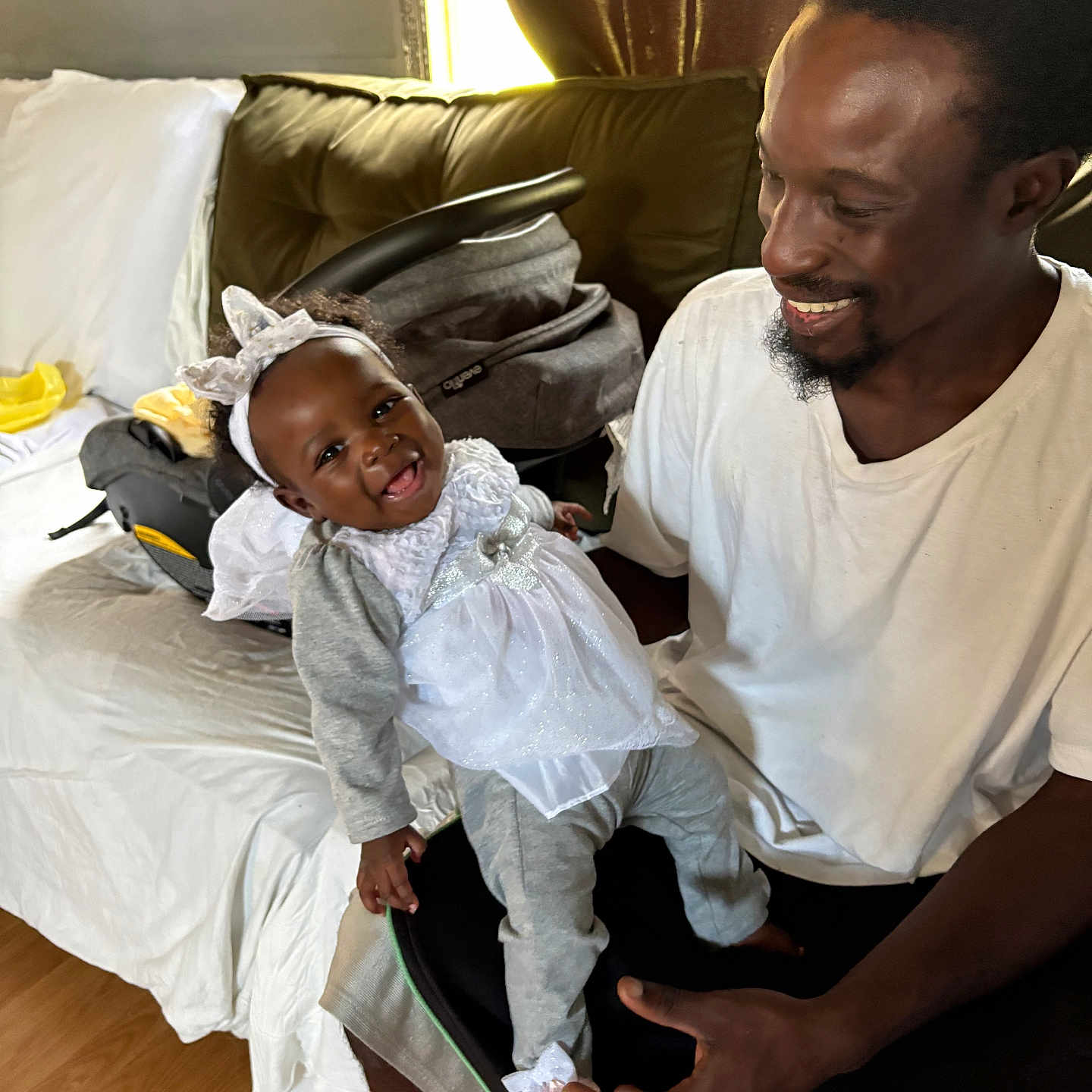 Annaliese joined the competition — help win amazing prizes! baby, child, man, smile, happy, indoor, living_room, couch, white_clothing, headband, baby_clothes, parent, family, holding, seated, window, curtains, car_seat, wooden_floor, casual