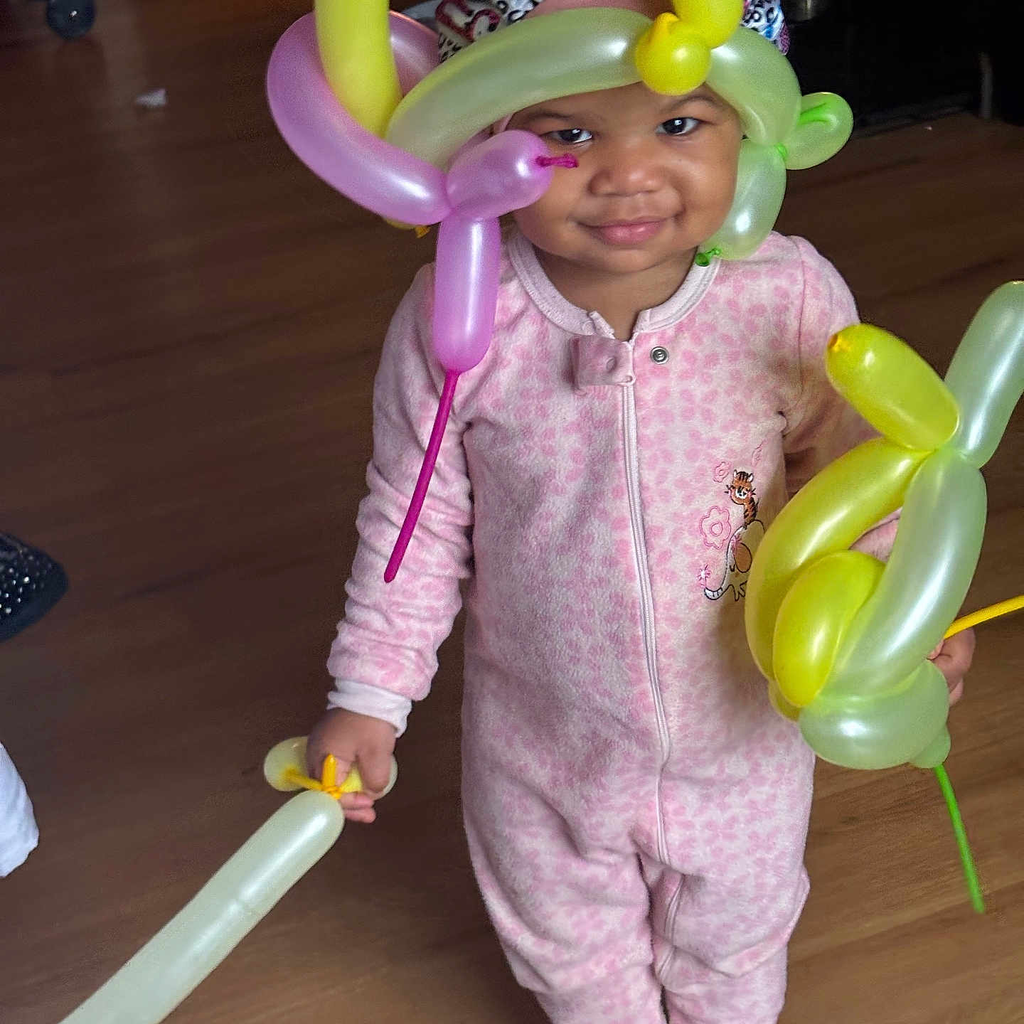 Ohana is registered to the contest to win money with this photo: baby, balloon, balloon_animal, balloon_hat, celebration, child, cute, footed_pajamas, happy, headwear, home, indoor, person, pink_onesie, playful, smile, standing, toddler, toy, wooden_floor