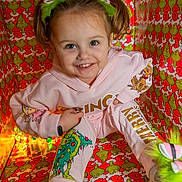 Violet is registered to the contest to win money with this photo: child, girl, smiling, pink_clothing, grinch, headband, pigtails, colorful, patterned_background, holiday_theme, sitting, box, happy, portrait, playful, bright_colors, fun, festive, indoor, footwear